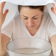 A woman with a towel over her head with her eyes closed, in front of a clear bowl of warm water and oil blends to promote better breathing