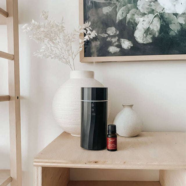 Aroma Wander portable diffuser in black and a Saje diffuser blend placed in front of two ceramic vases on a wooden table